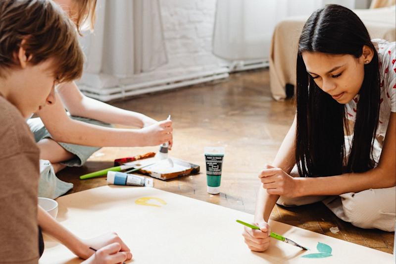 Three children paint with focus on large sheet of paper on floor in brightly lit room. None looking at camera.