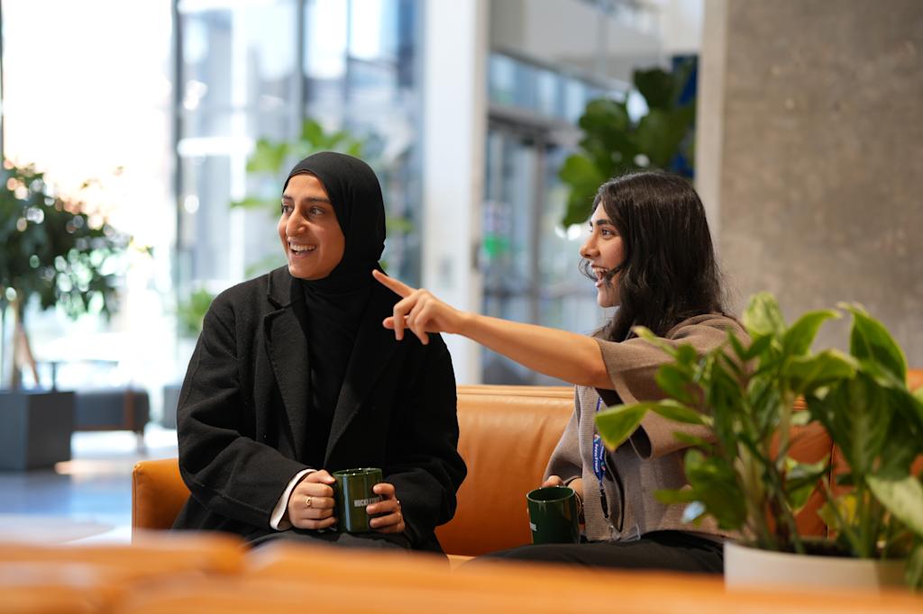 Two female researchers sit together on a sofa, each holding a mug.