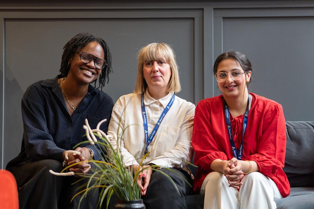 Three researcher interns sitting on a sofa in front of a grey wall. They are looking at the camera and smiling.