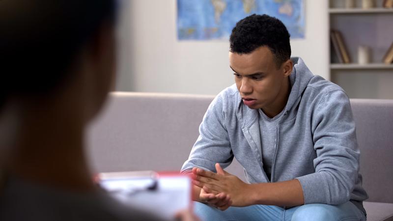 A teenage boy sitting in an office with an adult, looking down and rubbing his hands.