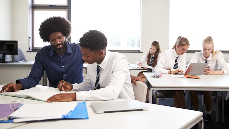 Secondary school teacher supporting a male student writing in a workbook at a classroom desk.