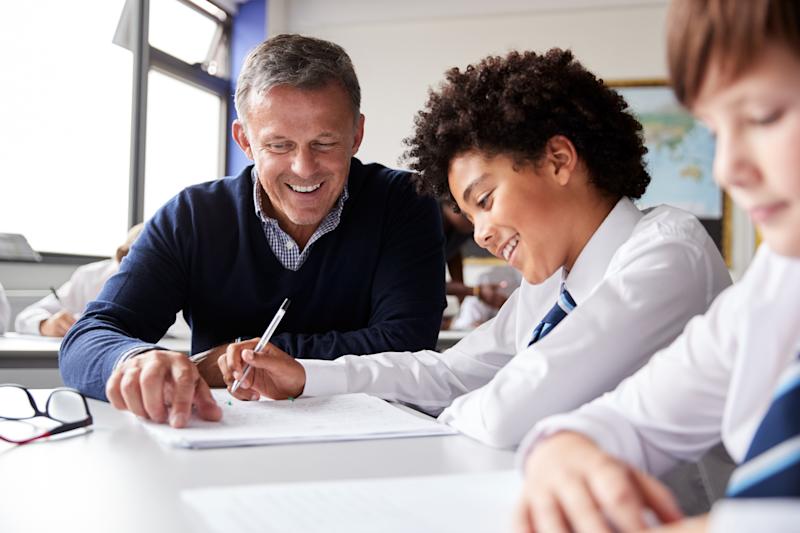 A male teacher sitting next to two secondary school children pointing at a textbook, smiling.