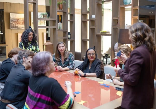 A group of training participants sitting around a table taking part in an activity. Two woman are standing - one at each end of the table.
