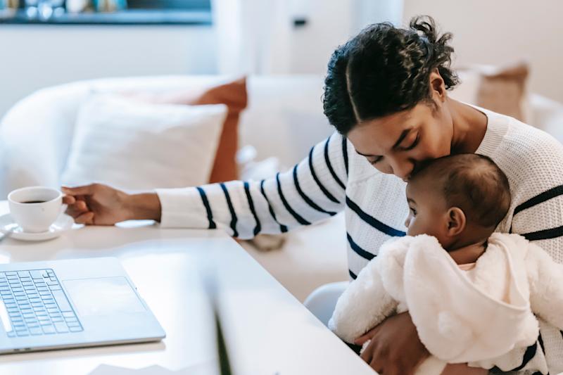 Woman kissing a baby's head as she holds them in one hand while the other holds a coffee cup, that sits on a table next to a laptop.