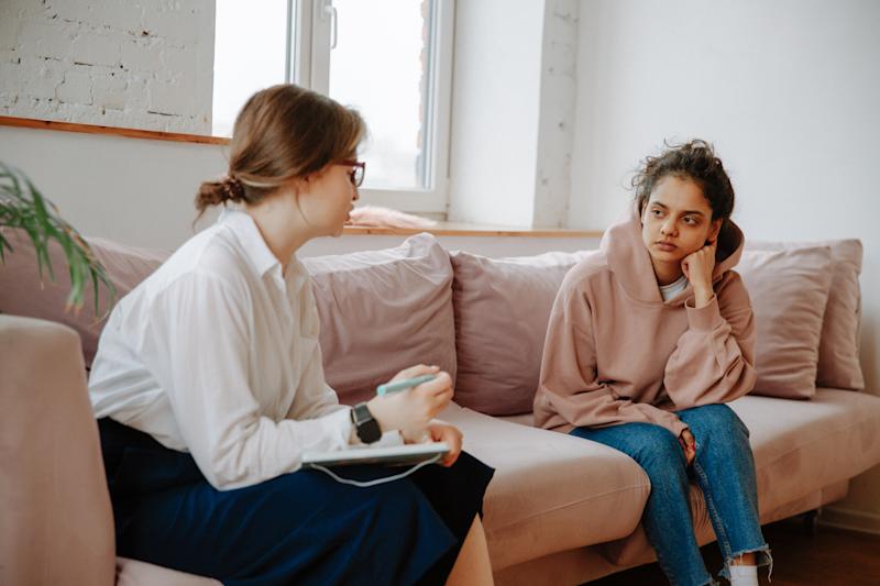 A young woman sitting on a sofa opposite a mental health practitioner  who is taking notes on a notepad.