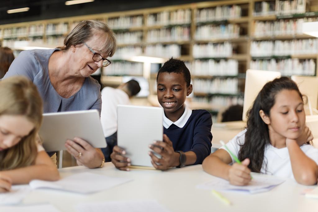 A photograph of a student in school uniform, using a tablet while a teacher looks on.