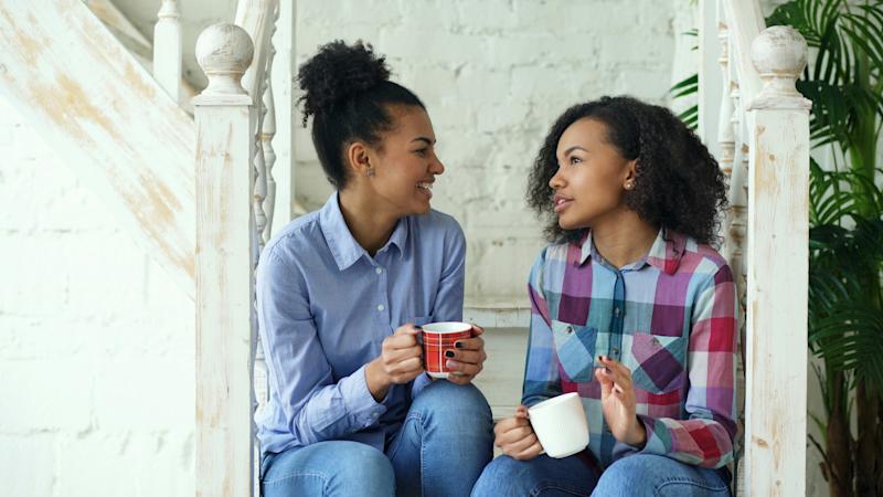 Two young women sitting on some white stairs in a house. They are holding mugs and talking.