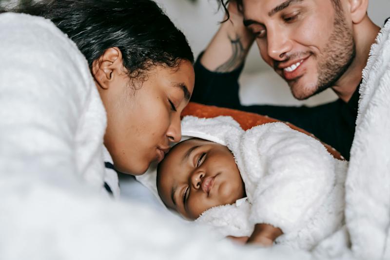A photograph of two parents cuddling their baby. The baby is fast asleep and the mother is kissing their forehead while the father watches on, smiling.