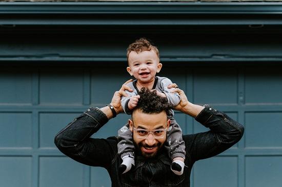 man with beard and glasses happily looks at camera while holding on to baby atop his shoulders.