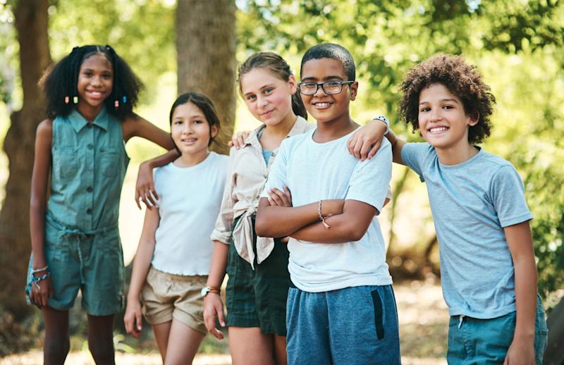 A group of children in their early teens stand and smile with their arms across each others shoulders. They are in a sunny forest.