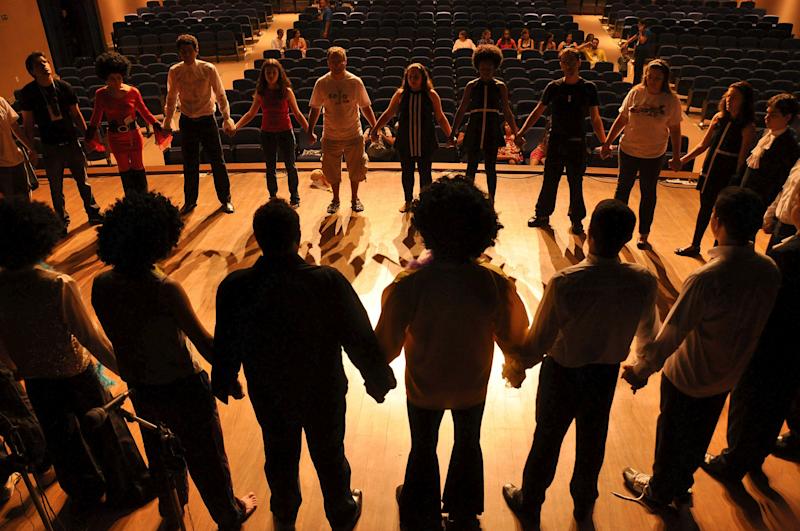 A group of young theatre students standing in a circle and holding hands on stage.