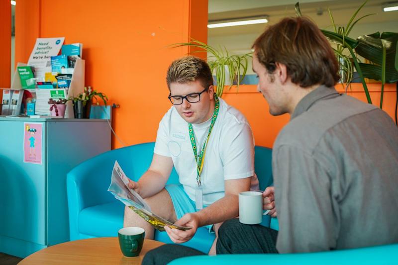 : A man wearing a sunflower lanyard sits looking at a magazine with another man holding a mug and looking at it.