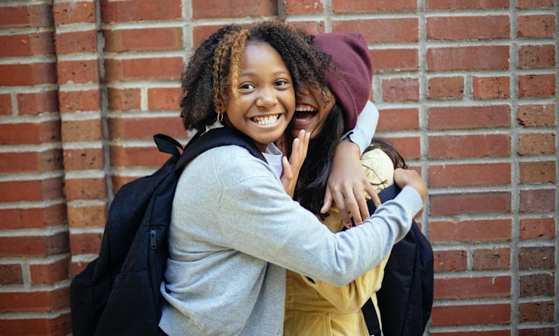 two children happily hug, one looking directly at camera. both have backpacks on. brickwork behind them.