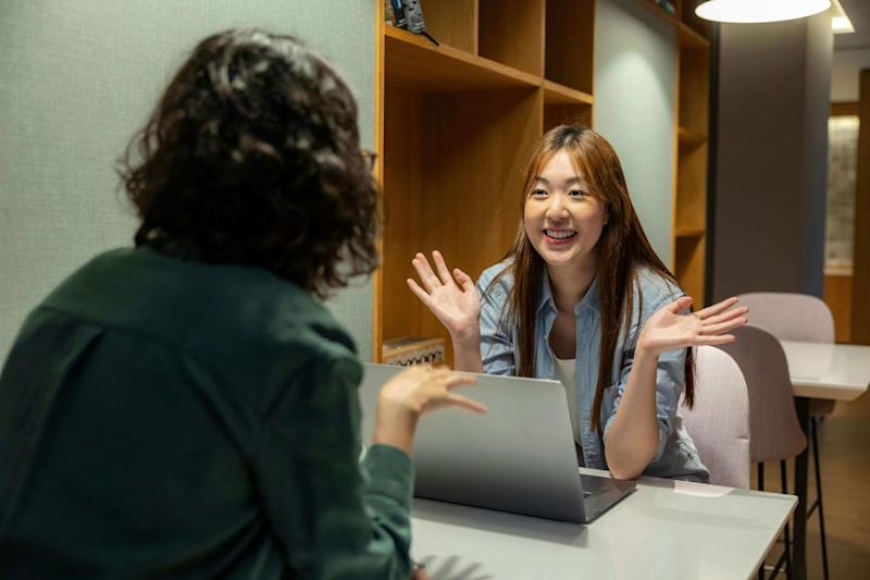 A photograph of two young people talking animatedly while sat at a table with a laptop between them.