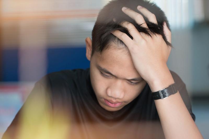 A teenage boy sits with his head in his hands as he looks downwards.