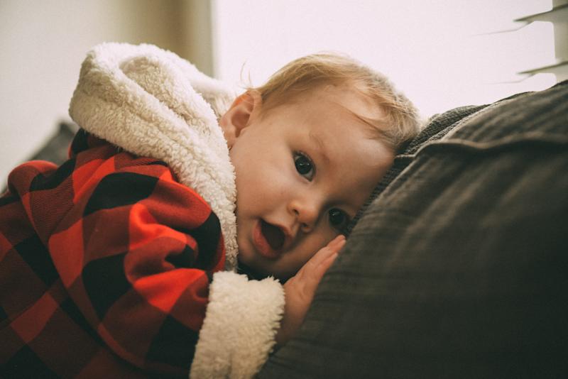toddler with face against sofa cushion looks at camera with mouth open