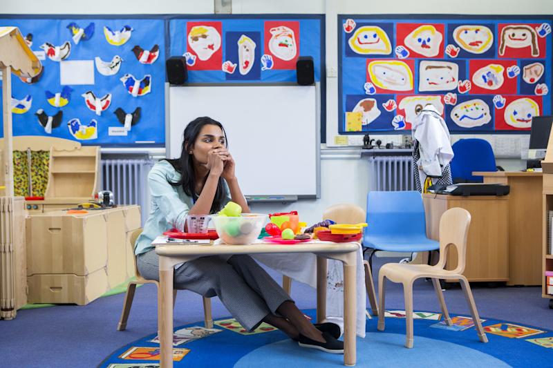 Stressed nursery or primary school teacher sitting at a table in a classroom, with hands covering her mouth and looking away from the camera.