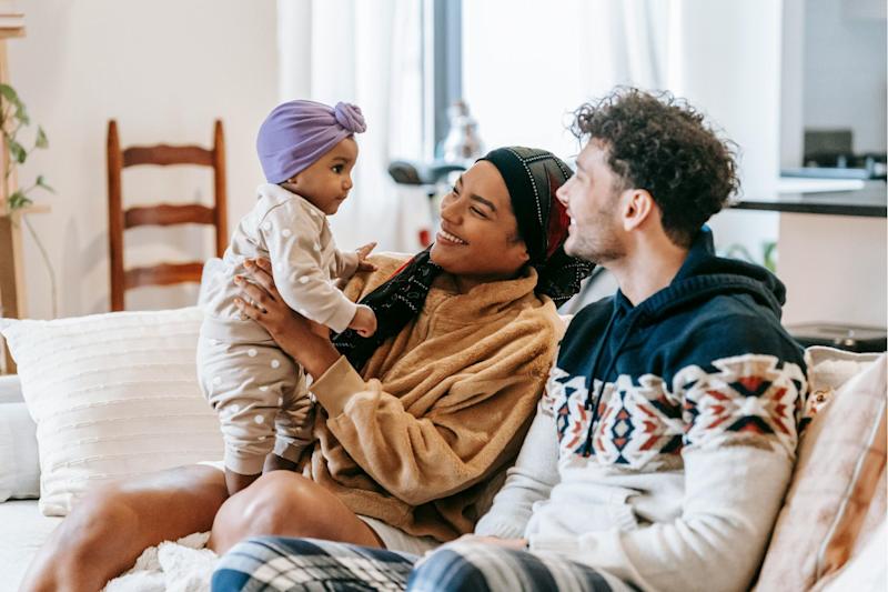 A mother and father sitting on a sofa at home. The mother is holding a young infant, and both parents are smiling at her.
