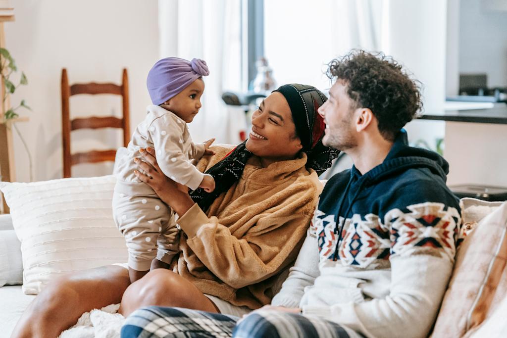 A mother and father sitting on a sofa at home. The mother is holding a young infant, and both parents are smiling at her.
