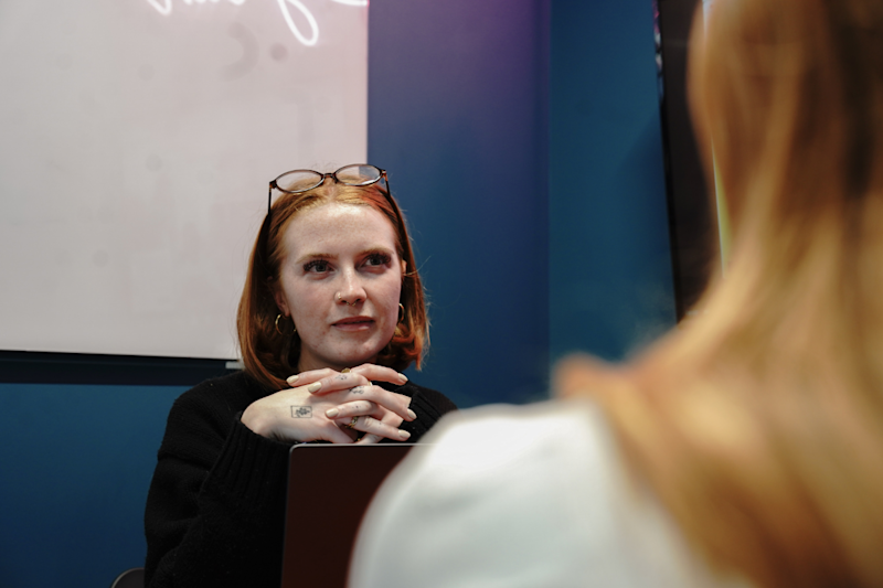 Young woman seated with her hands under her chin facing an interviewer.