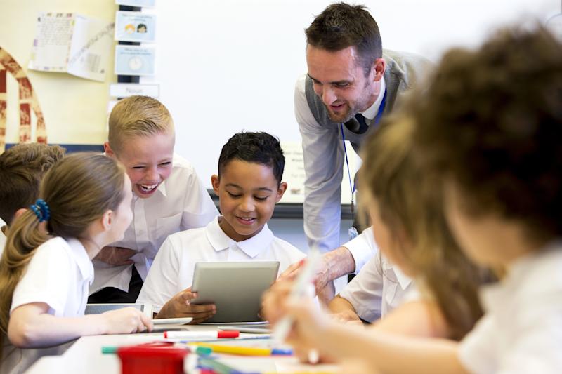 A smiling male teacher stands over supervising a group of happy children who are working on whiteboards and digital tablets.