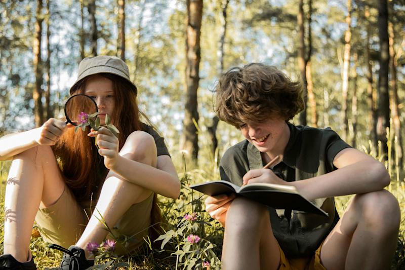 Two young people outdoors in nature. A girl uses a magnifying glass to closely examine a flower while a boy beside her smiles and writes in a notebook.