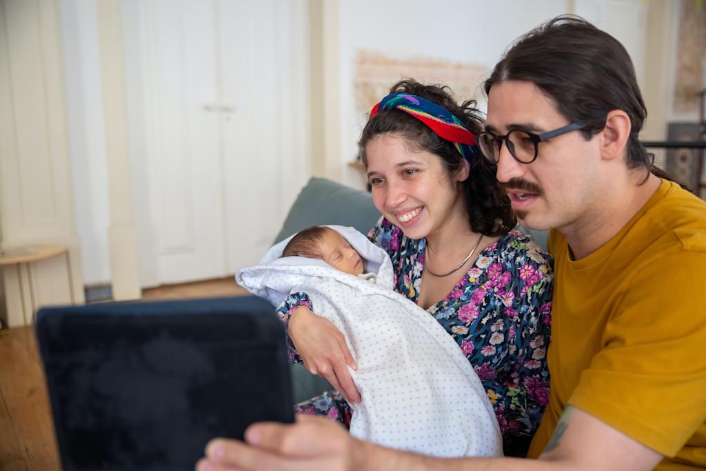 Mother holding newborn baby in arms next to father who is on a video call.