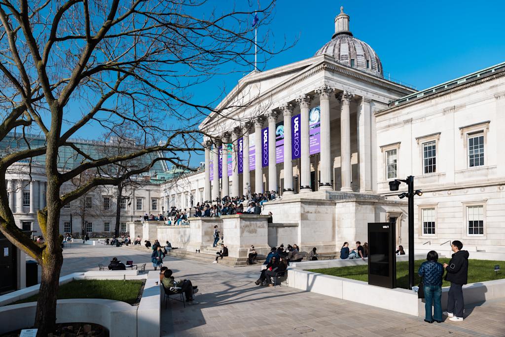 The UCL quad, a large sandstone building with columns and steps leading to the entrance, on a sunny day. Students are sitting on the steps and on benches outside.