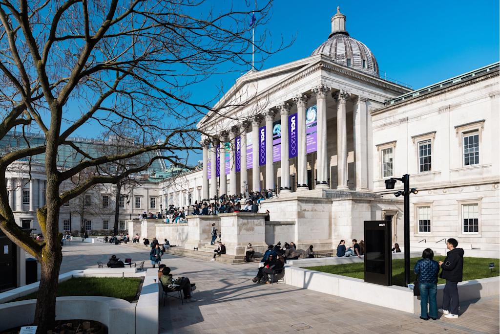 The UCL quad, a large sandstone building with columns and steps leading to the entrance, on a sunny day. Students are sitting on the steps and on benches outside.
