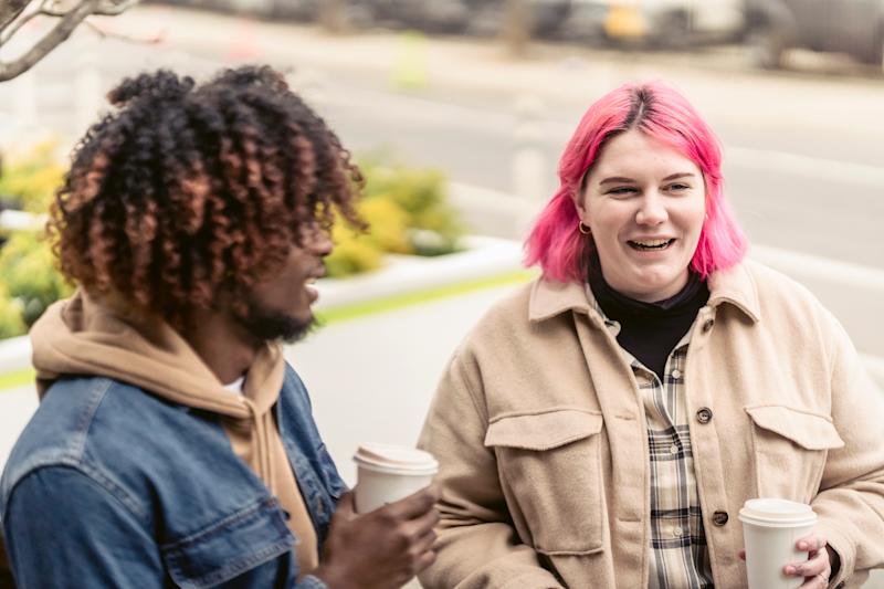 person with pink bob smiles holding coffee cup while man is to left blurred