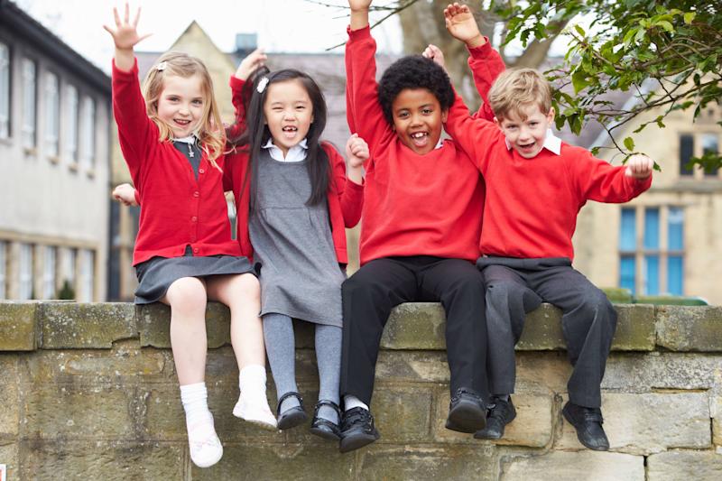 Four British primary school children wearing a red school uniform sitting on a wall smiling and raising their arms up in the air.