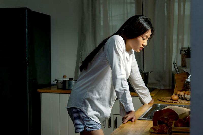 A teenage girl standing in a kitchen at night with her hands on the worktop, looking down and appearing upset.