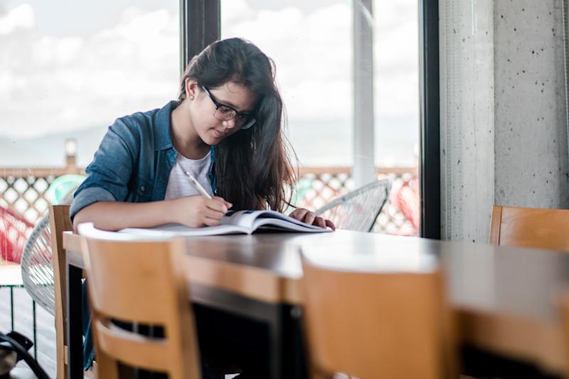 Female student sitting at a desk, writing in a textbook and looking down.