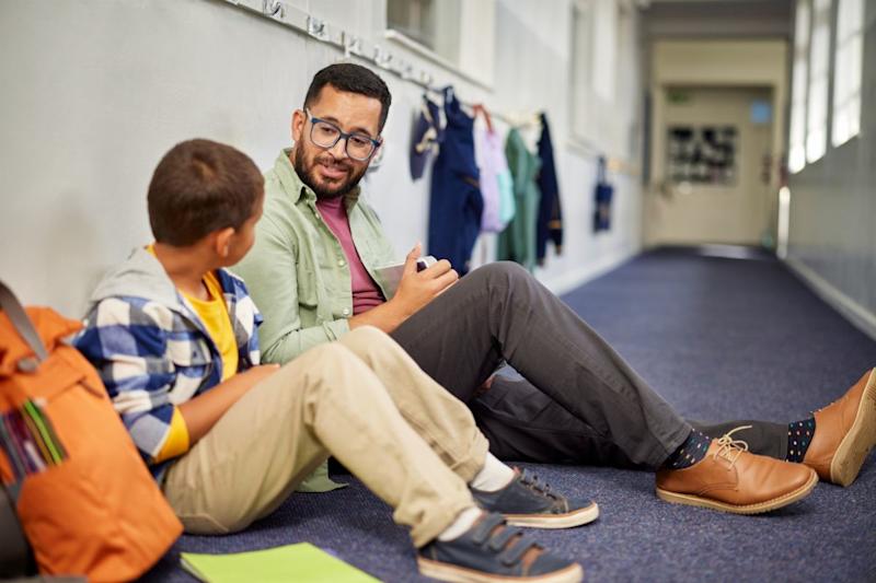 Man sitting on the floor of a corridor, smiling down at a young boy sitting beside him.