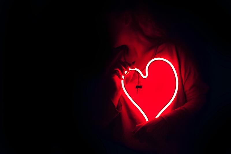 A photograph of a person surrounded by darkness, holding a neon red LED light in the shape of a heart.