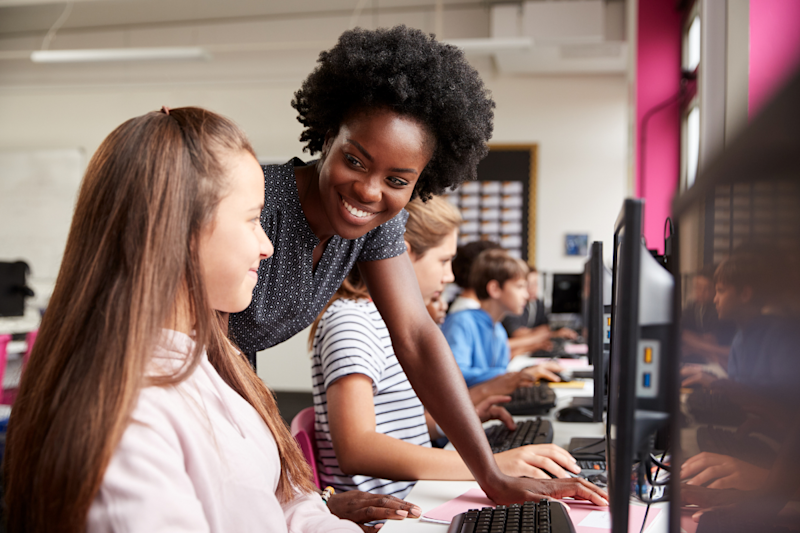 A teacher smiles as she leans over and helps a student working on a computer.