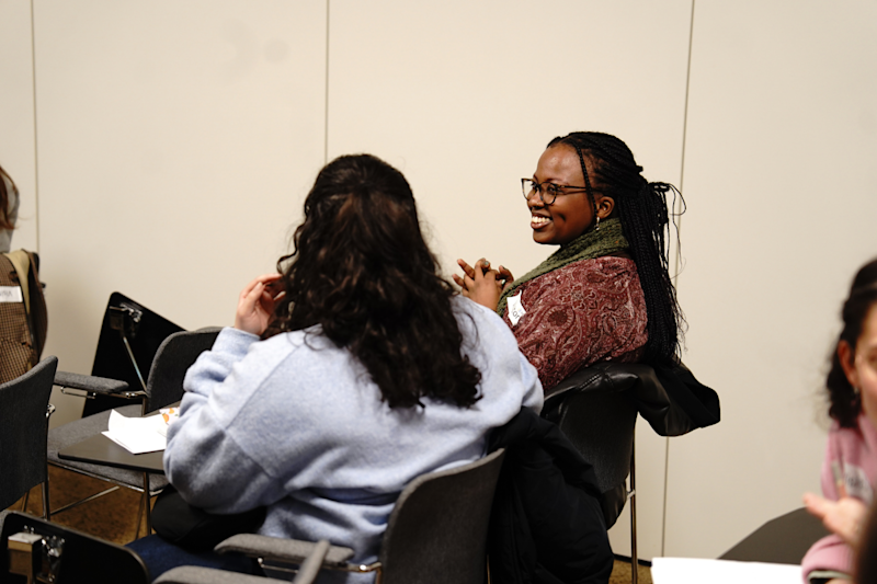 Two female students in a classroom, one smiling at the other while the second has their back to the camera.