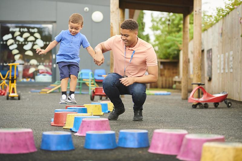 Nursery worker playing with a child in a playground while the child jumps and holds the man’s hand.