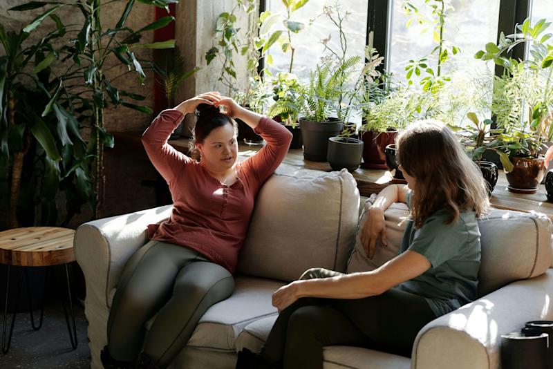 A young girl sitting on a sofa tying her hair up, seated opposite a woman, with plants visible on the windowsill in the background.