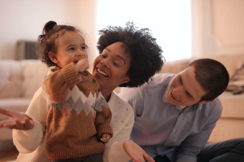 Man and woman looking happily towards a smiling young child. The child is close to the woman, whose hands are open with palms facing upwards.