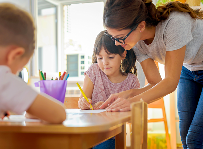 woman helps girl sat at table writing/drawing with yellow pencil. Girl concentrating, woman encourages, another child is working in foreground which is blurred