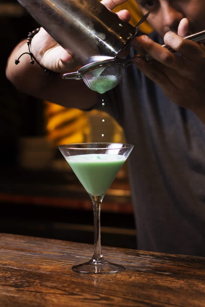 Bartender pouring a mint julep from a shaker through a strainer into a martini glass