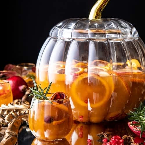 A glass of Thanksgiving sangria with fruit next to a pumpkin shaped punch bowl full of sangria