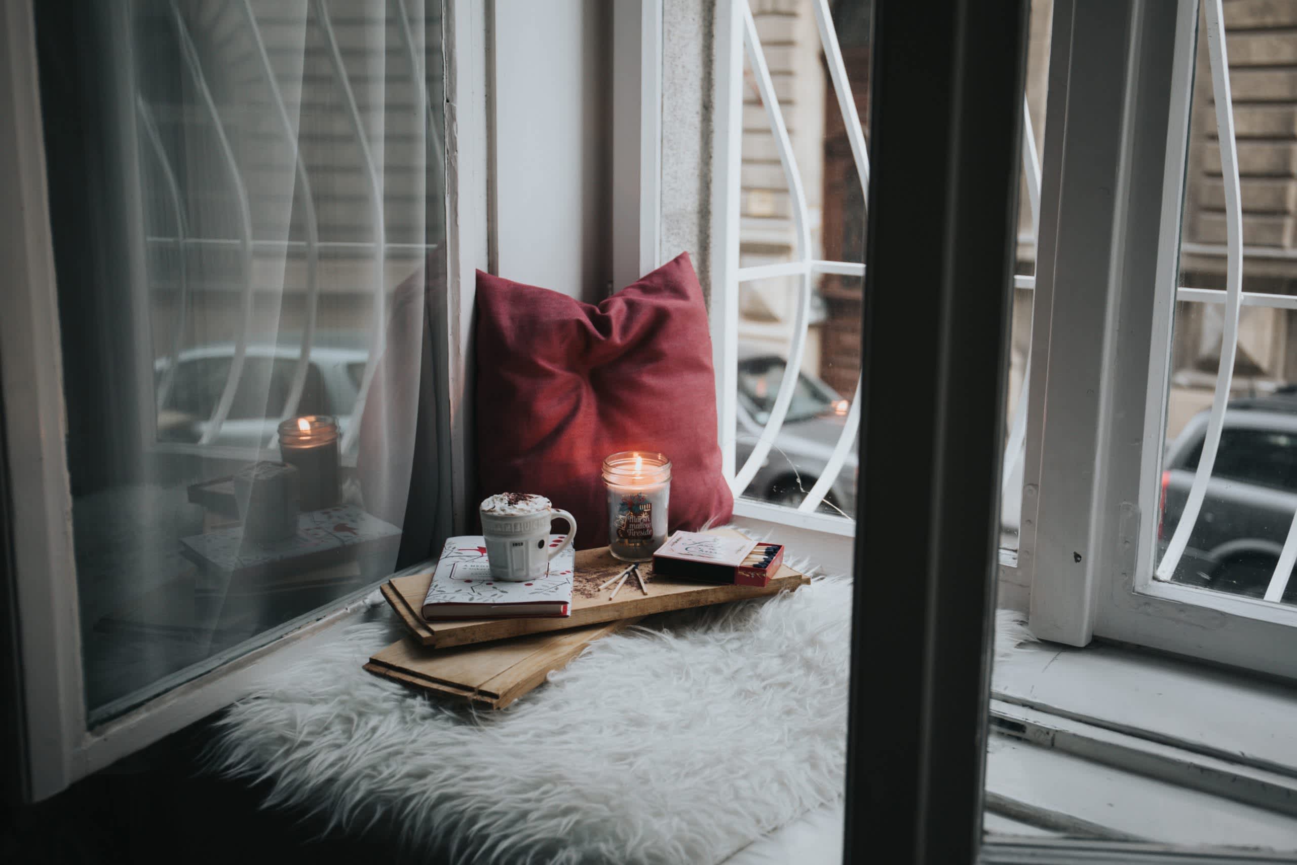 A spread of self-care items including cocoa, candles and a pillow by a window during winter