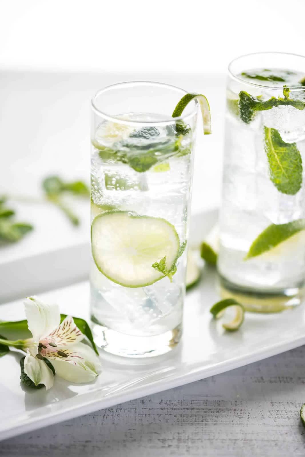 2 mojito mocktails in tall glasses with mint & lime on white tray with white flower  