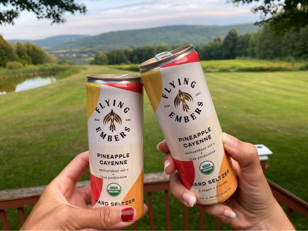 Two people cheersing with Flying Embers Pineapple Cayenne Hard Seltzer Cans