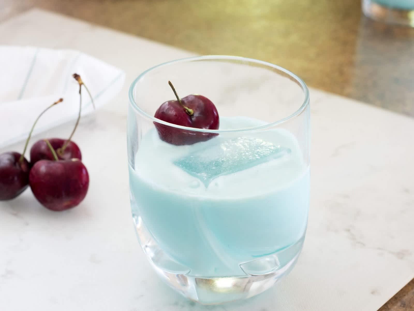 Rocks glass filled with baby blue frostbite cocktail & maraschino cherry 