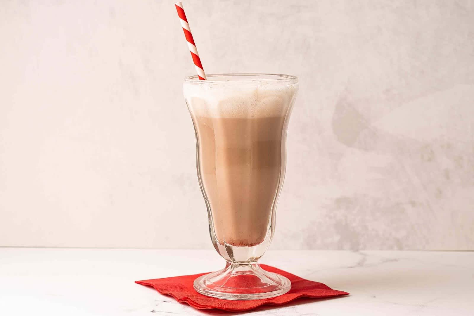 Chocolate egg cream in glass with striped straw on top of red napkin