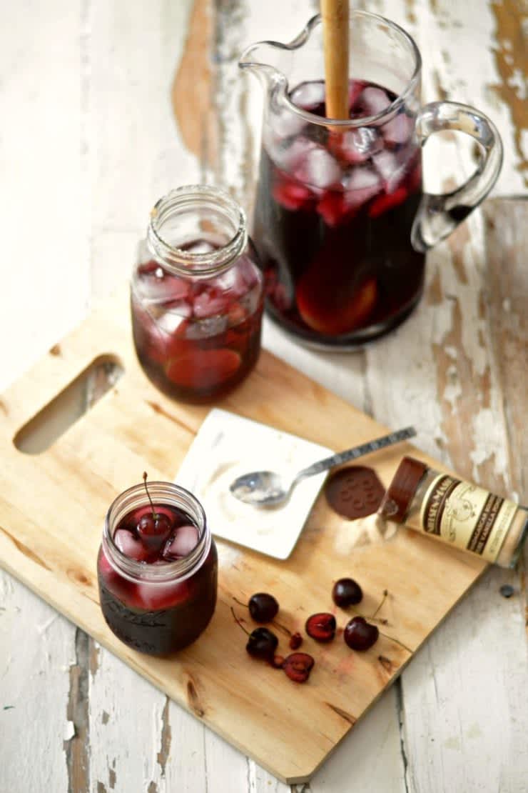 Two glasses of bourbon cherry vanilla sangria next to a pitcher on top of a cutting board