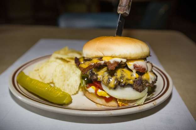 A bacon cheeseburger on a plate with a dill pickle spear and potato chips from Joe’s Tavern